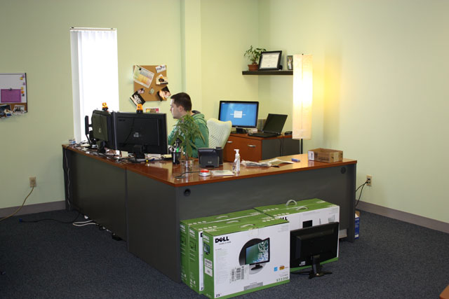 Man working at a desk in an office with dual monitors and boxed Dell computer monitors on floor.